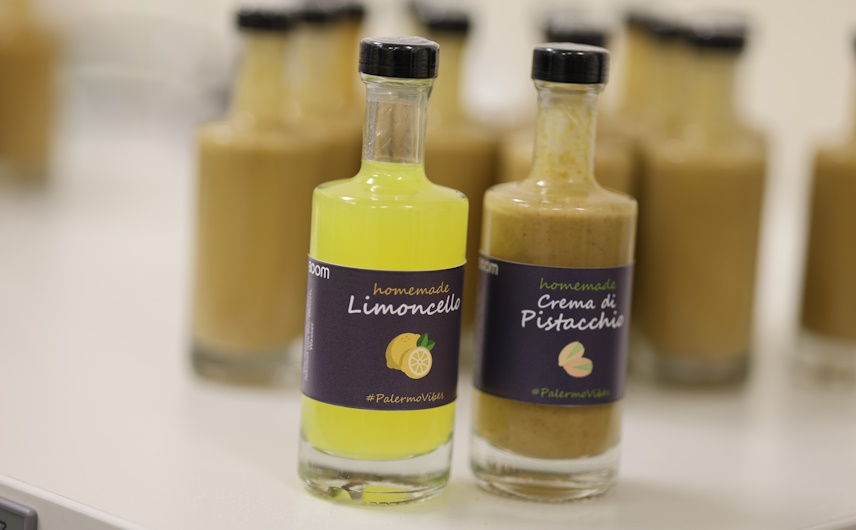 Eine Flasche mit gelbem Limoncello und eine Flasche mit bräunlicher Crema di Pistacchio stehen nebeneinander im Vordergrund des Bildes, im Hintergrund sind mehrere gefüllte Flaschen zu sehen. Sie sind das Mitarbeitergeschenk zu Weihnachten im Jahr 2024